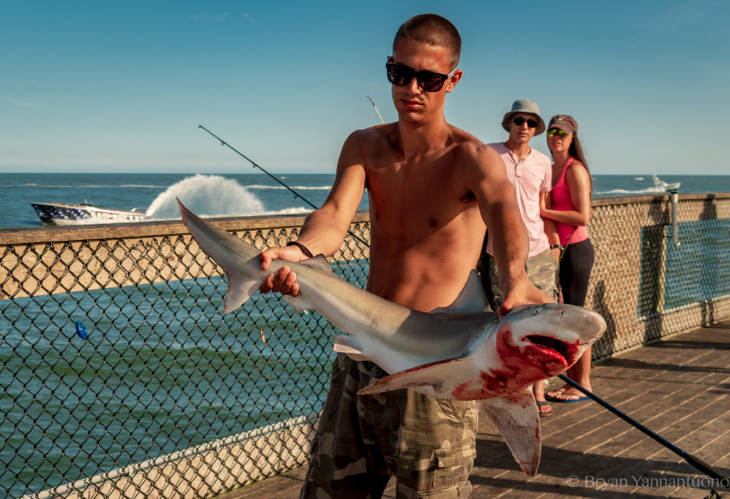 A shirtless man holds a bloody shark as a couple looks on in shock in the background