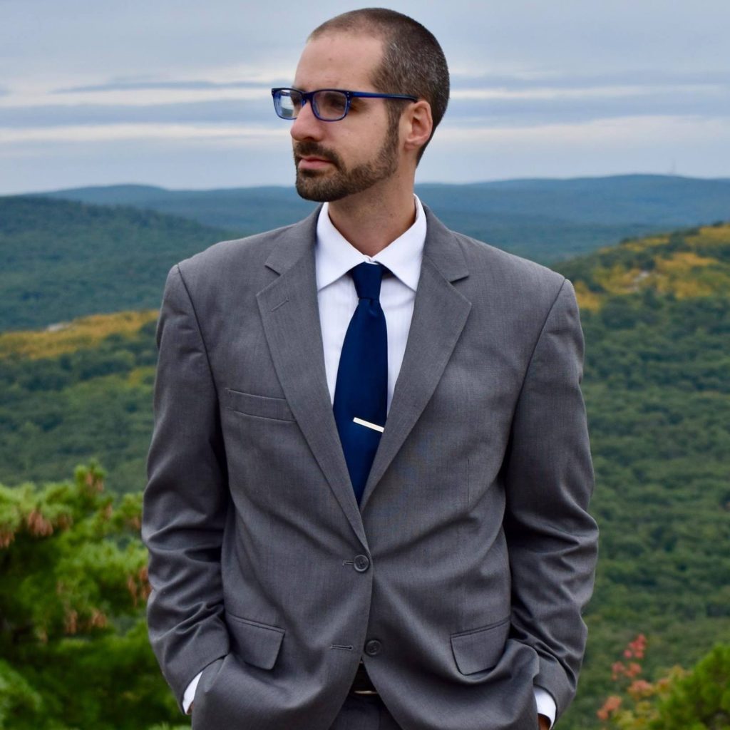 Bryan, a professional photographer in DC, stands in a dapper suit atop green mountains