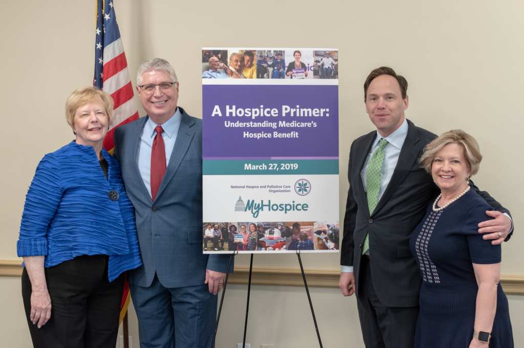 The photograph shows two men and two women standing in front of an American flag at a Capitol Hill Briefing